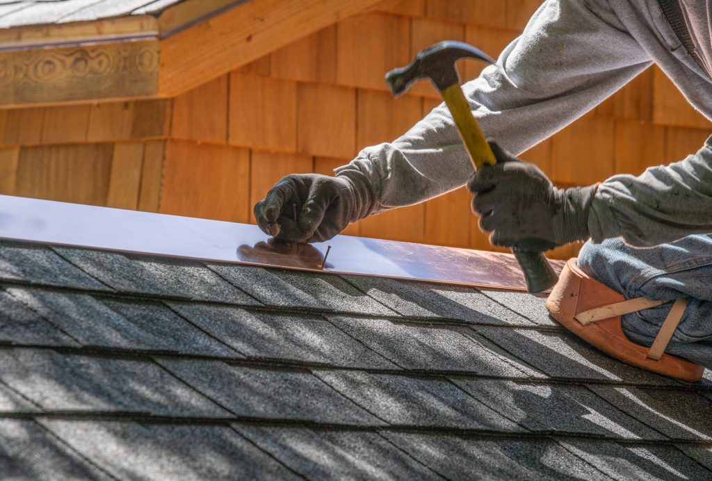 A roofing technician carefully hammers a nail through the edge of a shingle.